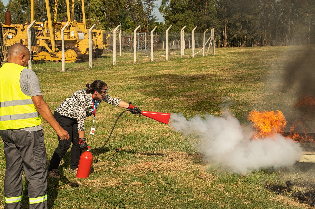 Woman in work attire using fire extinguisher to put out a practice fire in a field with a trainer